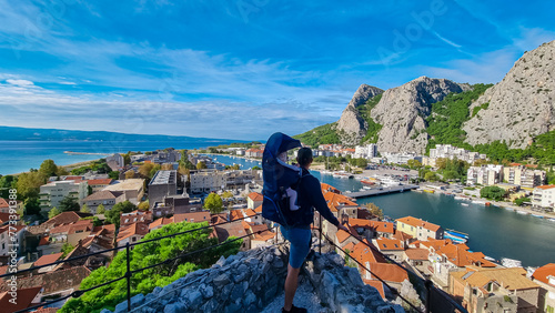 Father with baby carrier looking at coastal town Omis surrounded by Dinara mountains in Split-Dalmatia, Croatia, Europe. Majestic coastline of Omis Riviera at Adriatic Sea. Summer travel destination photo