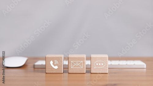 Three wooden blocks with contact icons for a phone, email, and chat. The blocks are arranged on a desk with a mouse and keyboard. Concept of communication and connectivity