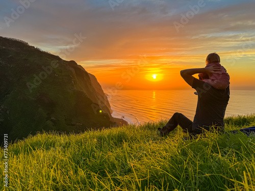 Toddler on shoulder of father on meadow with breathtaking sunset at viewing point Miradouro do Ponta da Ladeira  Madeira island  Portugal  Europe. Panoramic view of majestic coastline Atlantic Ocean