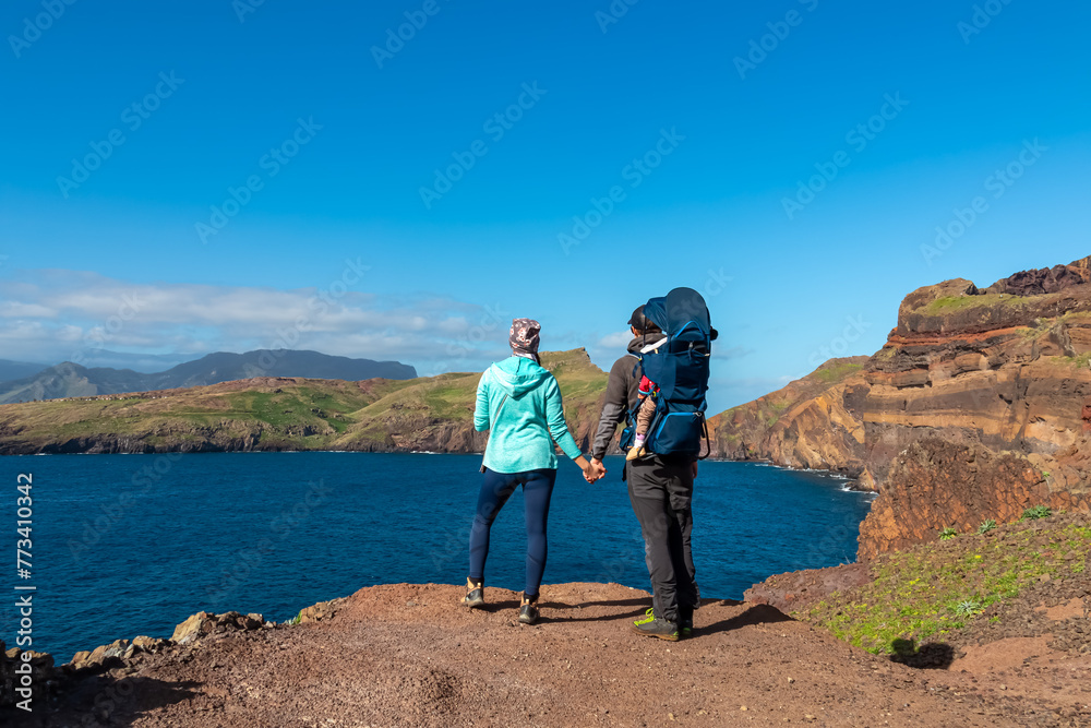Couple with baby carrier looking at majestic Atlantic Ocean coastline at Ponta de Sao Lourenco peninsula, Canical, Madeira island, Portugal, Europe. Coastal hiking trail along rocky rugged cliffs