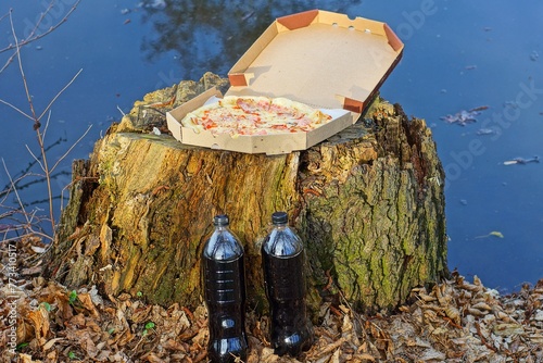 one round yellow fresh hot delicious pizza with sausage and cheese in a paper box and bottles of lemonade stands on an old wooden stump near the lake during the day on the street