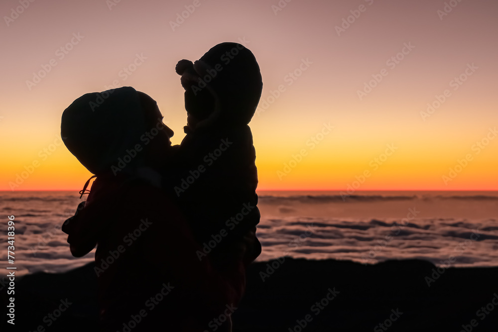 Silhouette of mother holding toddler and watching romantic sunrise on mountain peak Pico do Areeiro, Madeira island, Portugal, Europe. Panoramic view of clouds over the Atlantic Ocean. Orange red sky