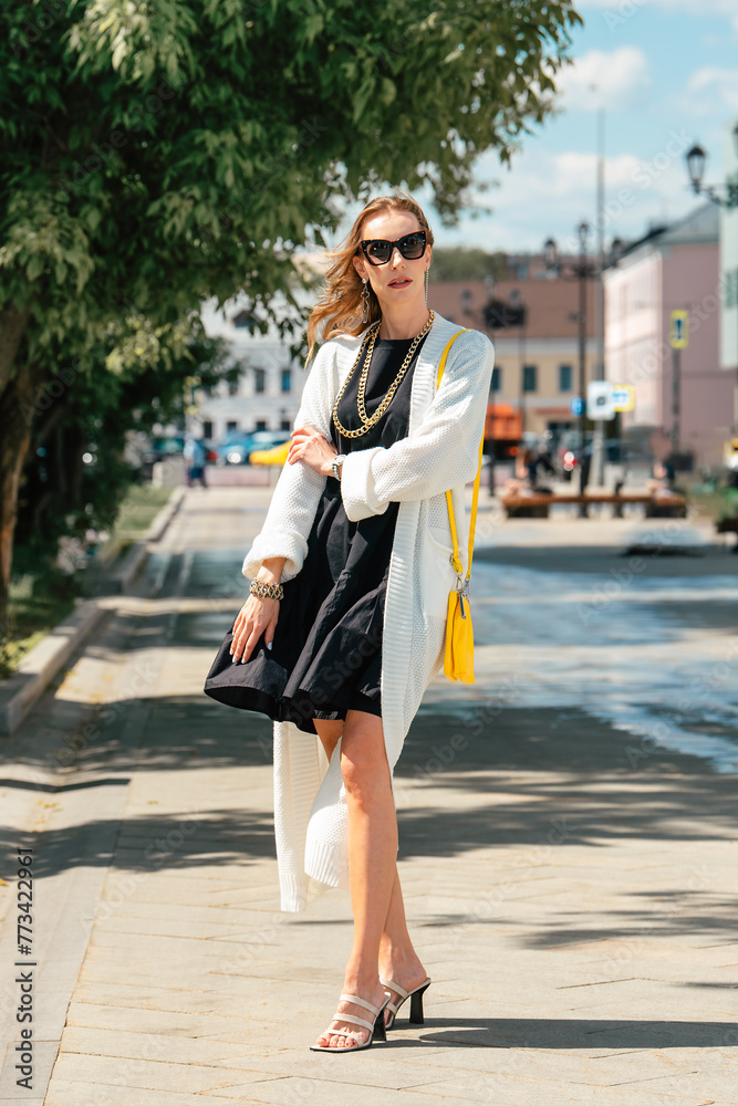 Stylish woman in a black dress and a white cardigan with a bright yellow handbag and a gold chain. A summer dress with frills flutters in the wind. Full length girl in the city in summer.