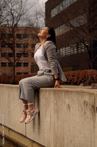 A young woman is casually perched on a concrete ledge, dressed in a stylish grey blazer, striped trousers, and pink sneakers, enjoying a sunny day in an urban setting.