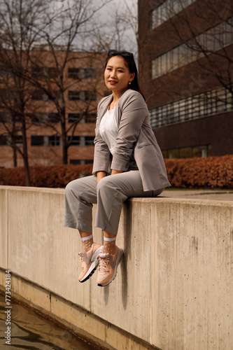 A young woman is casually perched on a concrete ledge, dressed in a stylish grey blazer, striped trousers, and pink sneakers, enjoying a sunny day in an urban setting.