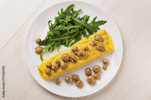 French three eggs omelette with mushrooms, arugula for a breakfast on a white plate on wooden background. Low carb breakfast. Top view.