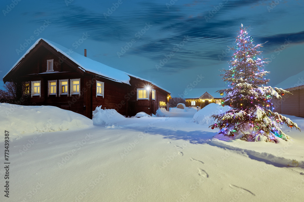 Naklejka premium Snowy night village in northern Europe on Christmas Eve, Christmas tree decorated with electric garlands stands on street in front of rural house.
