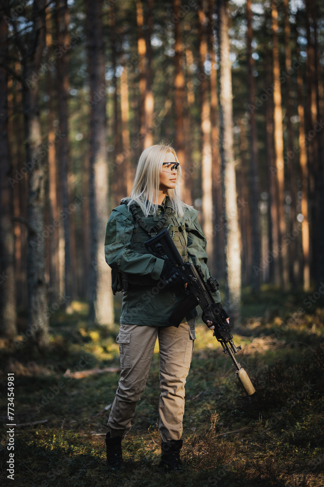 Beautiful military girl with a modern AK 12 suppressed rifle in the forest at war. Vertical photo.