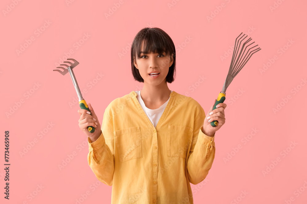 Beautiful young female gardener with gardening rakes on pink background Beautiful young female gardener with gardening rakes on pink background