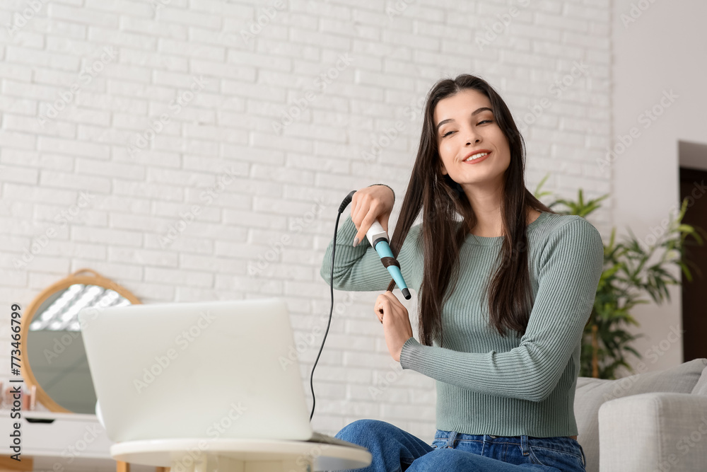 Beautiful young happy female blogger with laptop and curling iron recording video at home Beautiful young happy female blogger with laptop and curling iron recording video at home