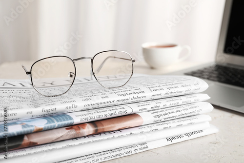 Stack of newspapers with eyeglasses, laptop and cup of tea on white table in office