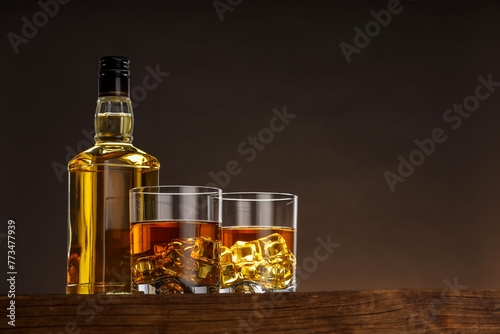 Whiskey with ice cubes in glasses and bottle on wooden table against brown background, space for text © New Africa
