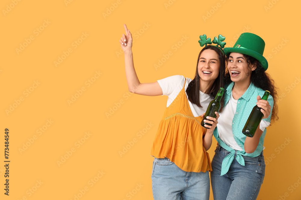 Beautiful young women with bottles of beer pointing at something on yellow background. St. Patrick's Day celebration