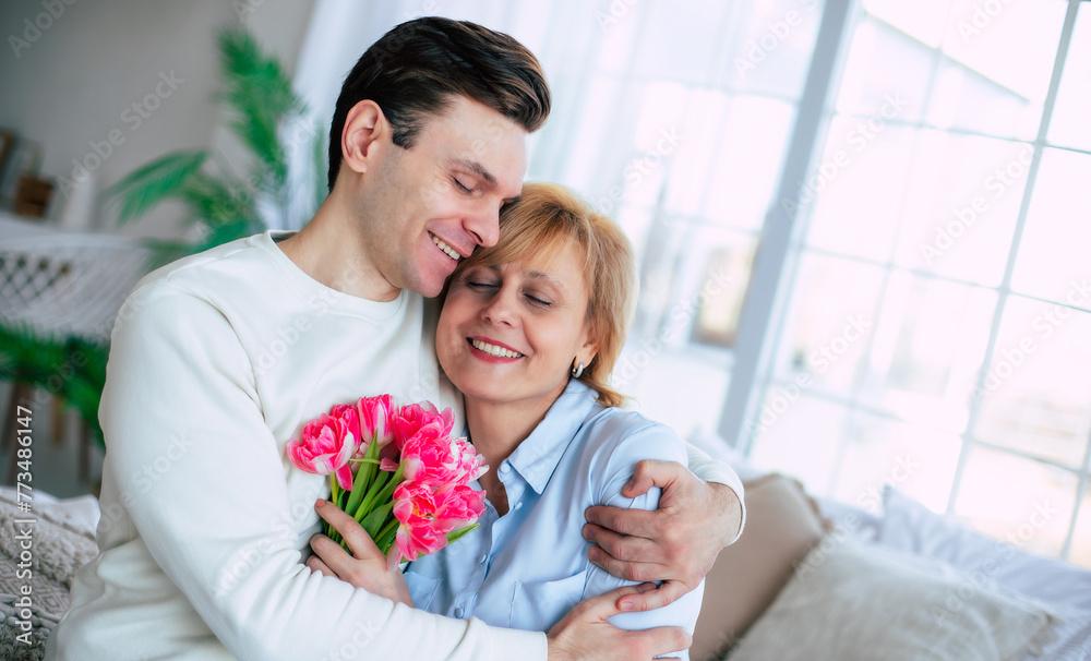 Close up photo of smiling son embracing his cute mother at home with bouquet of tulips. Birthday, Mother's day, women day, retired. Close up photo of smiling son embracing his cute mother at home with bouquet of tulips. Birthday, Mother's day, women day, retired.