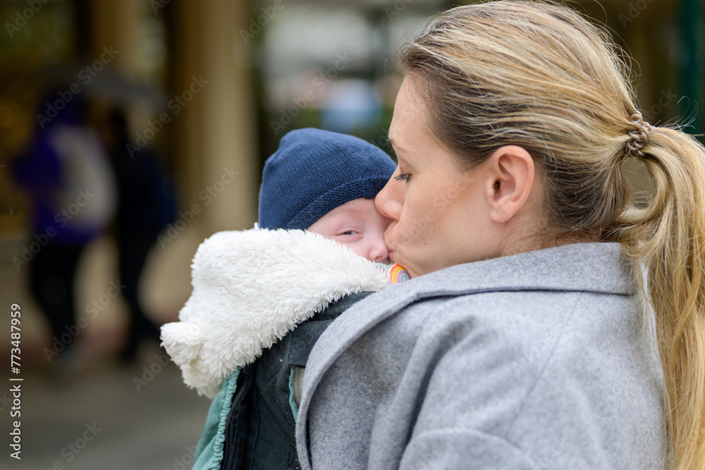 Late-term mother in her 40s lovingly holding her newborn baby in a baby carrier and kissing him gently Late-term mother in her 40s lovingly holding her newborn baby in a baby carrier and kissing him gently