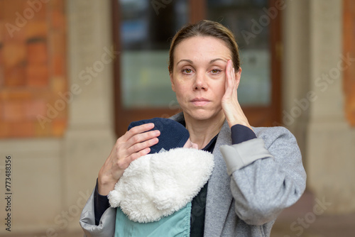 Exhausted and stressed late-term mother in her 40s holding her newborn baby in a baby carrier and looking annoyed at the camera