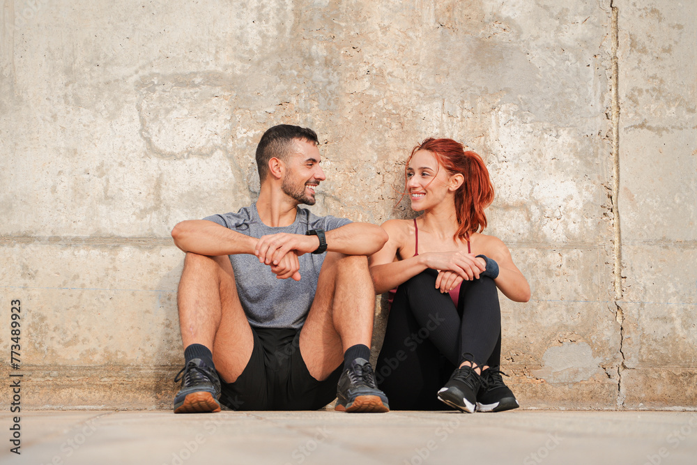 Sport couple taking a break of the workout. Tired young people looking each other, sitting on the floor, recovering energy and having a conversation. Coach man and athletic woman talking and resting