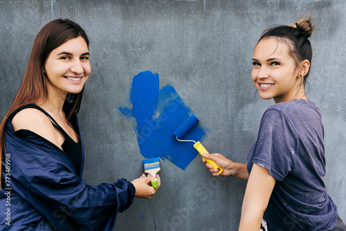 Painting work, smiling young women paint gray wall with blue paint using brush and roller. photo