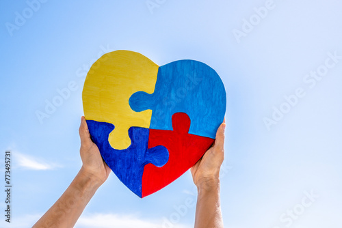 World autism awareness day, Autism spectrum disorder concept. Adult and child hands holding together colorful painted puzzle heart on blue sky background