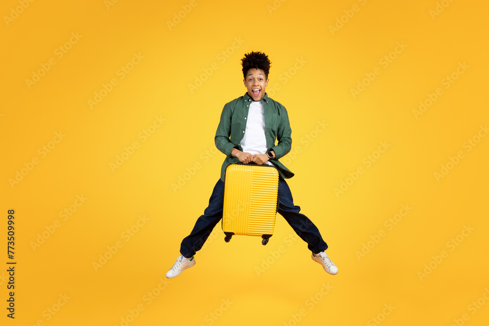 Excited traveler african american guy jumping with yellow suitcase
