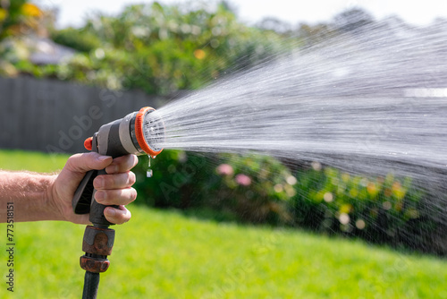Garden hose with nozzle. Man's hand holding spray gun and watering plants, spraying water on grass in backyard.