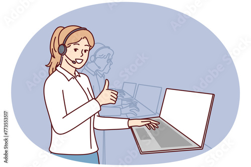Woman in headphones for call center stands near laptop and looks at camera showing thumbs up. Girl employee of psychological support service or software user consultation smiling