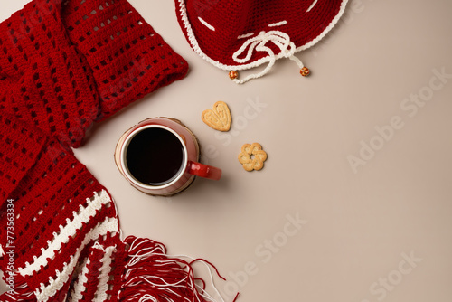 Knitted scarf and hat with mug of tea and cookies on beige background