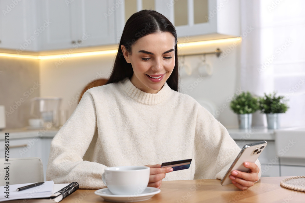 Happy young woman with smartphone and credit card shopping online at wooden table in kitchen Happy young woman with smartphone and credit card shopping online at wooden table in kitchen