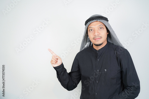 Cheerful Asian Muslim man wearing Arab turban sorban pointing hand finger at empty space on isolated photo