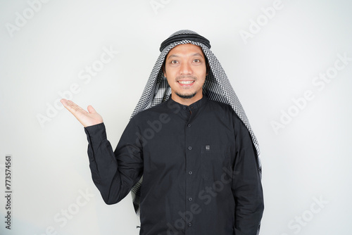Cheerful Asian Muslim man wearing Arab turban sorban pointing hand finger at empty space on isolated photo