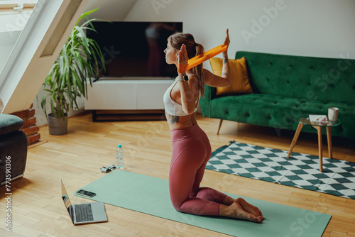 A woman is kneeling on a yoga mat in a plantfilled room, using an orange resistance band photo