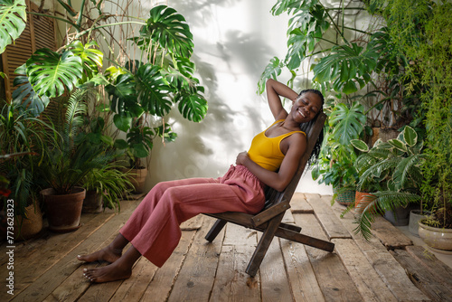 Joyful African American woman with smile reflecting enjoyment of much-needed break sits on chair in sunlit ambiance of urban jungle room. Pleased black female resting in home lounge with houseplants. photo