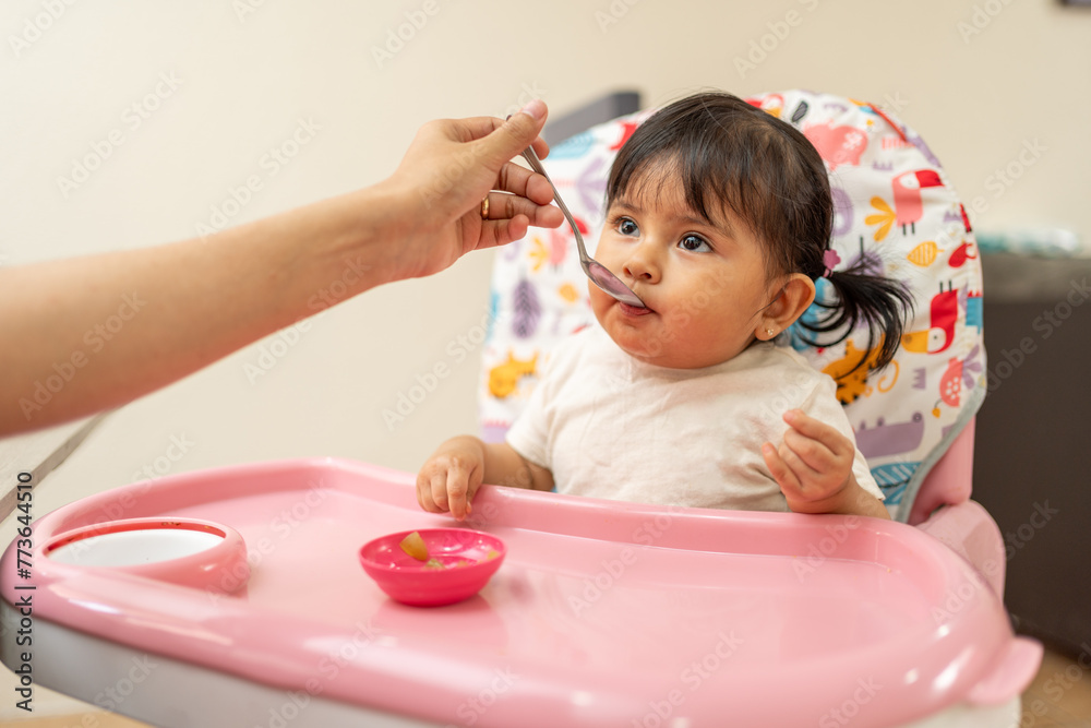 portrait of a Hispanic baby girl eating, hand of Latin mother feeding her toddler in a baby high chair portrait of a Hispanic baby girl eating, hand of Latin mother feeding her toddler in a baby high chair