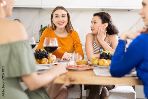 Cheerful female friends enjoying home gathering with wine and light snacks at kitchen table, engaging in lively conversations..