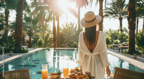 A woman in white dress and straw hat stands at the table with food by poolside of luxury hotel resort