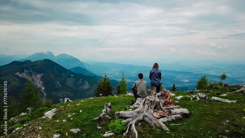 Hiker couple on alpine meadow near Feistritzer Spitze (Hochpetzen) with panoramic view of majestic mountain peaks Karawanks and Julian Alps, Carinthia, border Austria Slovenia. Hiking trail Petzen