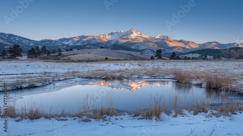 A tranquil winter scene featuring a partially frozen pond  golden sunlit snow-capped mountains  and a clear blue sky at dawn.