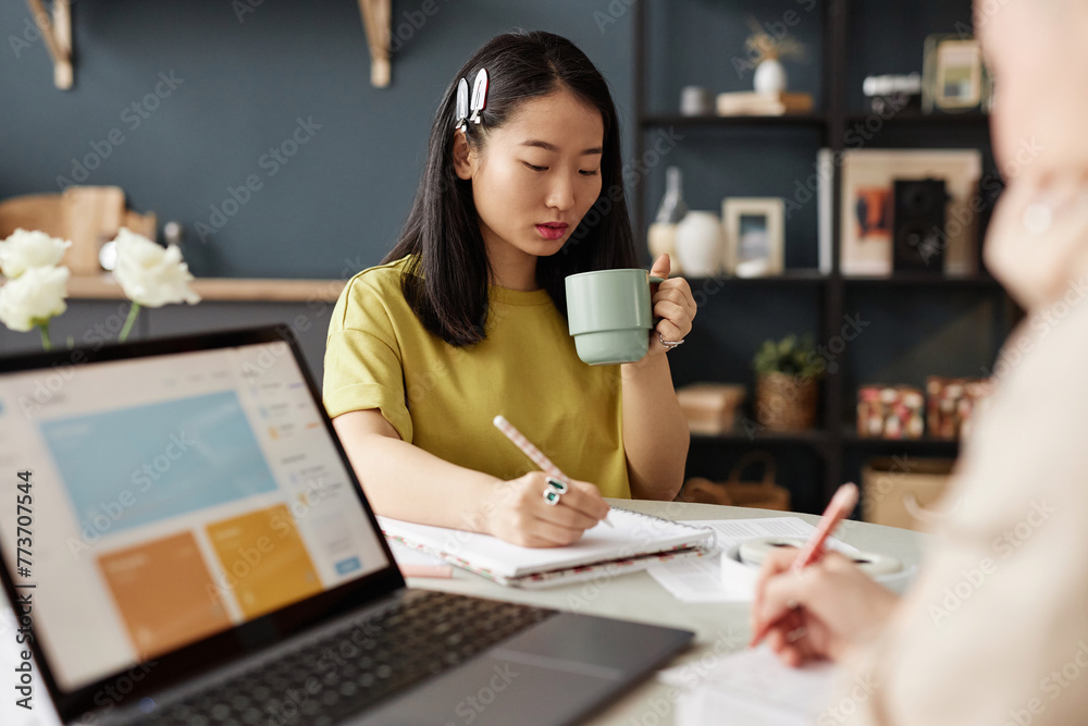 Selective focus shot of young adult Asian woman holding cup of tea or coffee discussing school project with her friend while making notes Selective focus shot of young adult Asian woman holding cup of tea or coffee discussing school project with her friend while making notes