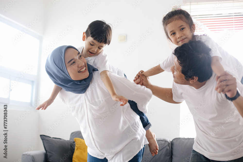 Obraz premium Cheerful Asian mom and dad piggy back their children in living room