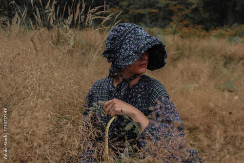 Pioneer woman in a dark blue dress sitting in a grassy meadow. Foraging basket. Historical reenactment. photo