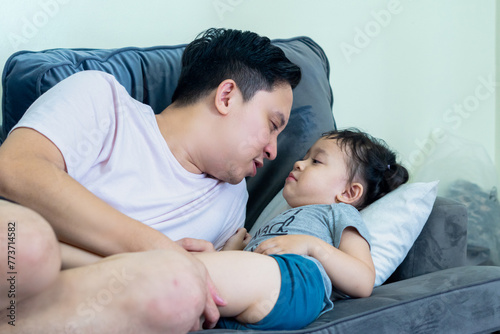 Asian man lying on sofa in living room with little girl on top, both smiling, father and daughter relationship. photo