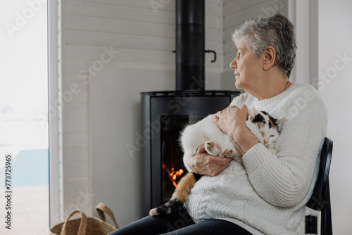 an elderly woman dressed in a comfortable home sweater sits by a lit fireplace with a fluffy cat in her arms