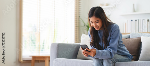 Happy young asian woman relax on comfortable couch at home, typing chat message on smartphone, smiling girl use cell phone chatting, searching information on browser wireless internet, online shopping photo