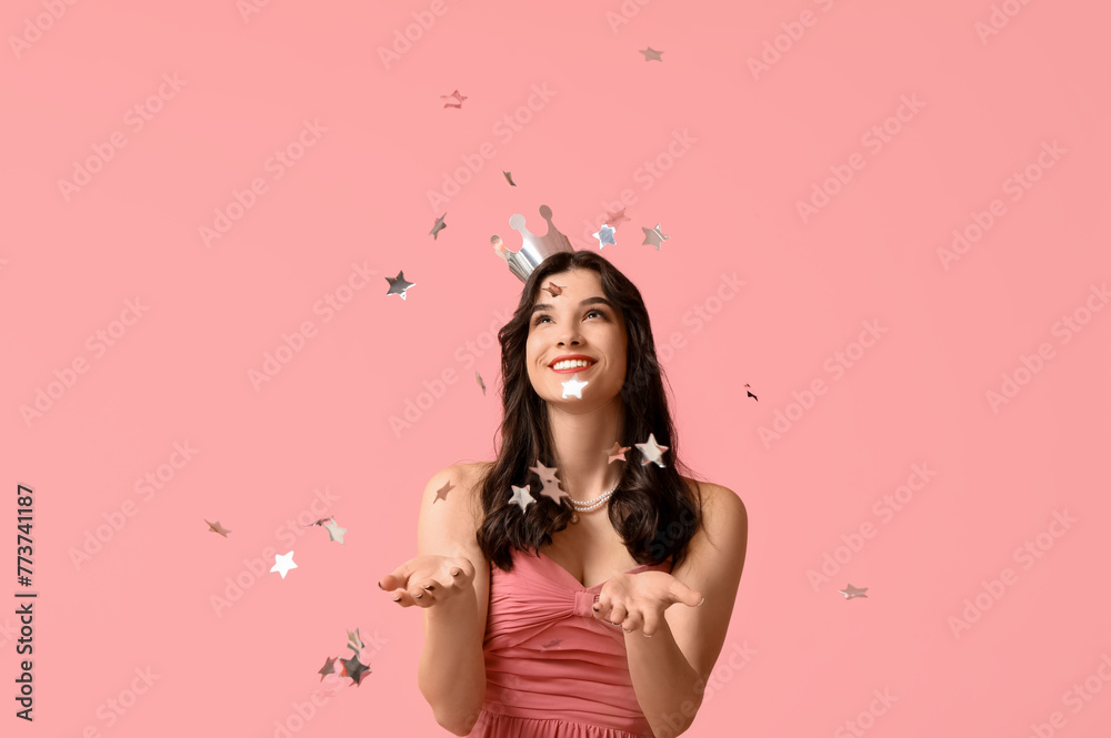 Beautiful young woman in stylish prom dress and paper crown with confetti on pink background