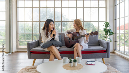 Two friend sit on sofa in a bright living room with large glass wall. Play guitar and use smartphone as microphone, sing happily. Tablet computer, coffee cup and decorated pot plant are on the table.
