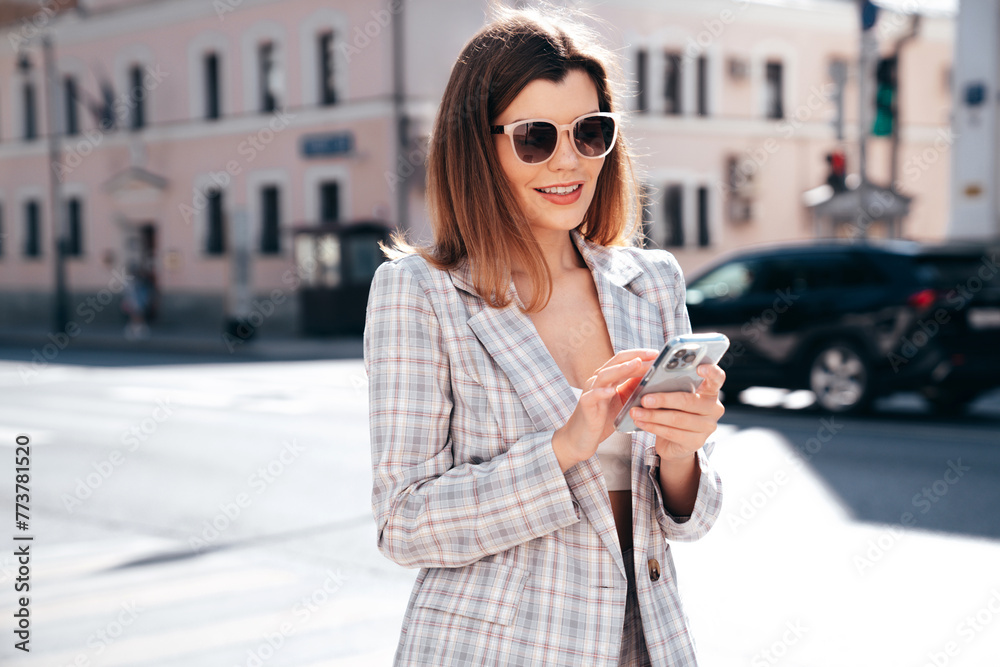 Obraz premium Young beautiful woman in nice trendy white jacket suit. Sexy smiling model posing in street. Fashionable female outdoors. Cheerful and happy. Holds smartphone, uses phone apps, looks at screen