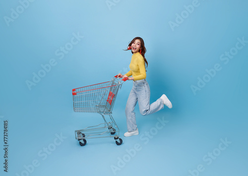 Full length happy Asian teen woman pushing empty shopping cart or shopping trolley running promotion supermarket isolated on blue background.