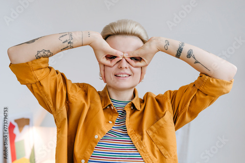 Smiling woman holding fingers to eyes like glasses near white wall photo