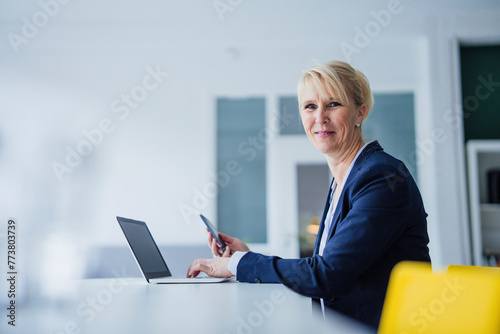 Smiling businesswoman sitting with smart phone and laptop at desk photo