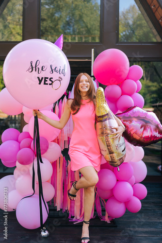 Vertical portrait of loving redhead bride in summer wedding dress posing holding various colorful festive foil helium balloons standing by country house, closed eyes with happy expression.